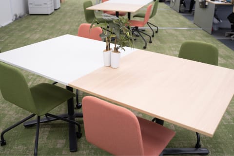 緑のカーペットの上に配置された木目調のテーブルと、グリーンとピンクのオフィスチェア。テーブルの上には観葉植物が置かれている。