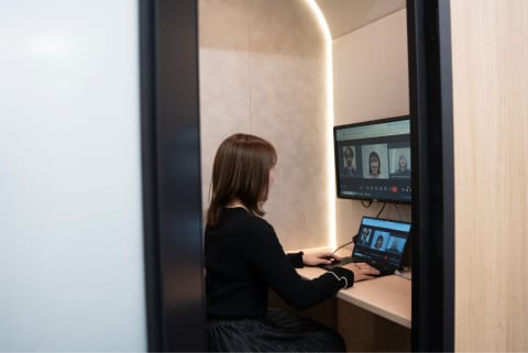 個別ブース内でビデオ会議をしている女性。モニターとノートパソコンに複数の人物の顔が映っている。