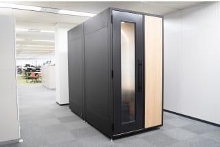 オフィスフロアに設置された黒と木目調のWeb会議用ブース。ガラス扉付きの個室型デザインで、奥にはオープンスペースのオフィスが見える。